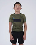 A young boy wearing an olive green athletic rash guard with a black chest panel and black shorts, standing against a light gray studio background.