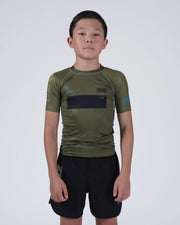 A young boy wearing an olive green athletic rash guard with a black chest panel and black shorts, standing against a light gray studio background.